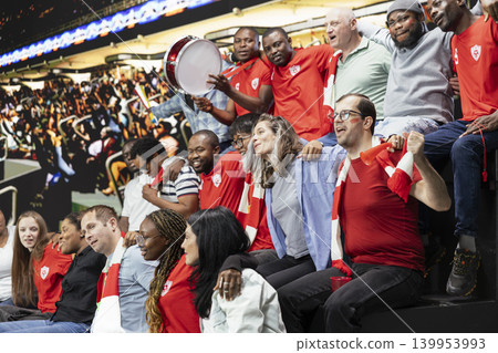 Packed stadium crowd of diverse soccer fans cheering with red scarves, drums and vuvuzelas during an exciting live football match. Fans cheering and showing team support, unity. Packed stadium crowd of diverse soccer fans cheering with red scarves, drums and vuvuzelas during an exciting live football match. Fans cheering and showing team support, unity. 139953993