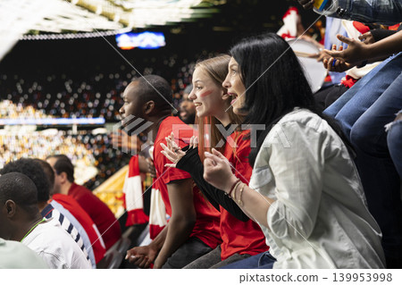 Excited female soccer fans leaning forward and cheering from stadium seats with diverse supporter crowd, red shirts and scarves during live match action. Worried supporters focus on the game. Excited female soccer fans leaning forward and cheering from stadium seats with diverse supporter crowd, red shirts and scarves during live match action. Worried supporters focus on the game. 139953998