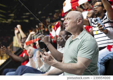 Soccer fan using phone to monitor in-play live betting odds and bet ticket during football match at the stadium. Adult man supporter tracking bet slips and live sportsbook updates. Soccer fan using phone to monitor in-play live betting odds and bet ticket during football match at the stadium. Adult man supporter tracking bet slips and live sportsbook updates. 139954000