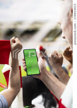 Vertical mockup of football supporter holding phone with green screen in packed stadium during live soccer match. Fans cheering in the stands with authentic sports emotion, supporter culture. Vertical mockup of football supporter holding phone with green screen in packed stadium during live soccer match. Fans cheering in the stands with authentic sports emotion, supporter culture. 139954015