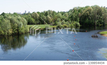 Silent Riverbank Bordered By Abundant Verdant Foliage And Floating Signs. Peaceful Lakeside Panorama Showcasing Still Waters Mimicking Natural Habitat With Lush Trees And Floating Markers 139954434