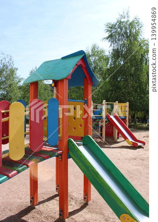Colorful children's playground equipment featuring slides, climbing structures, and play panels in a sunny outdoor setting surrounded by trees and blue sky Colorful children's playground equipment featuring slides, climbing structures, and play panels in a sunny outdoor setting surrounded by trees and blue sky 139954869