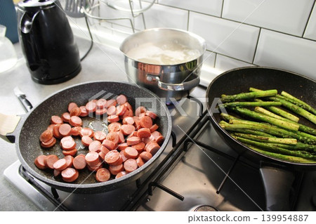 Cooking process with sliced sausages in a frying pan, asparagus in another pan, and boiling water in a pot on a modern kitchen stove Cooking process with sliced sausages in a frying pan, asparagus in another pan, and boiling water in a pot on a modern kitchen stove 139954887
