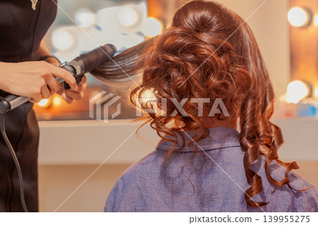 Hairdresser styles hair with blow dryer while client sits in salon chair facing mirror and lights. Concept of hair care, salon service, beauty preparation, hairstyle creation workflow Hairdresser styles hair with blow dryer while client sits in salon chair facing mirror and lights. Concept of hair care, salon service, beauty preparation, hairstyle creation workflow 139955275