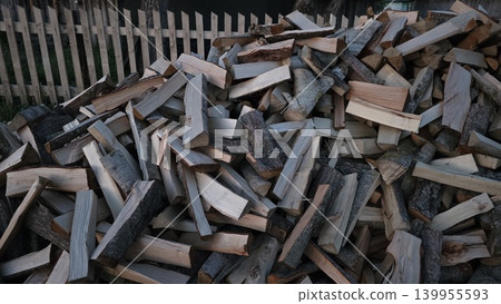 Large pile of chopped firewood lying outdoors near wooden fence in countryside, concept of preparation for winter heating season Large pile of chopped firewood lying outdoors near wooden fence in countryside, concept of preparation for winter heating season 139955593