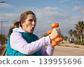 Woman exercising with orange dumbbells shows effort expression during workout along coastal promenade. Outdoor fitness training captures exertion moment with both arms raised and mouth open exhaling. 139955696