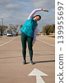 Woman 45 years old stretching sideways raises one arm overhead during sport workout outdoors in parking lot near sea. Outdoor exercise session focuses on lateral flexibility and warm up on sunny day. 139955697