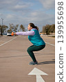Woman doing squat exercise holds orange dumbbells extended forward during sport workout outdoors in parking lot. Side view of workout taking place near sea on sunny day in open air setting. 139955698
