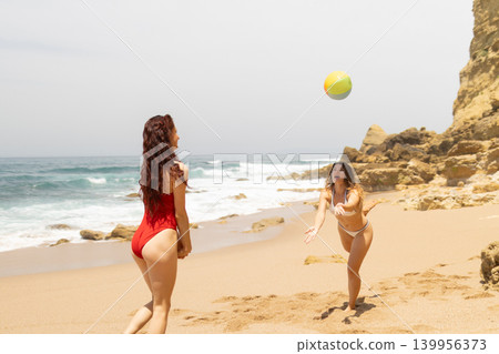 Two women playing beach volleyball with a colorful ball on a sandy shore, ocean waves crashing in the background under a clear sky Two women playing beach volleyball with a colorful ball on a sandy shore, ocean waves crashing in the background under a clear sky 139956373