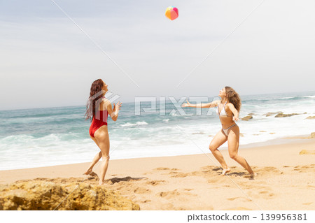 Two women playing with a colorful beach ball on sandy shore near ocean waves, wearing swimsuits under bright sunlight with rocky formations in the background 139956381