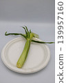 A single, freshly cut young corn (baby corn) with the husk still on, placed on a white plate. 139957160