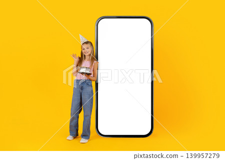 Smiling little girl standing beside oversized smartphone mockup with birthday cake Smiling little girl standing beside oversized smartphone mockup with birthday cake 139957279
