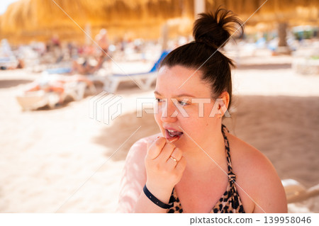 Caucasian woman applies lipstick while relaxing on a beach in Sharm El Sheikh Egypt during sunny summer vacation. Beach resort lifestyle personal care and leisure tourism on the Red Sea Caucasian woman applies lipstick while relaxing on a beach in Sharm El Sheikh Egypt during sunny summer vacation. Beach resort lifestyle personal care and leisure tourism on the Red Sea 139958046