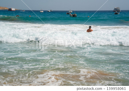 Caucasian man swims in sea waves near boats on the Red Sea coast in Sharm El Sheikh Egypt with turquoise water and summer beach weather. Beach resort lifestyle and seaside tourism on the Red Sea Caucasian man swims in sea waves near boats on the Red Sea coast in Sharm El Sheikh Egypt with turquoise water and summer beach weather. Beach resort lifestyle and seaside tourism on the Red Sea 139958061