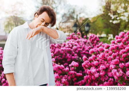 Sick man outdoors suffering from spring allergy and hayfever. He is sneezing into his elbow due to pollen from the bright pink blooms and flowers. Sick man outdoors suffering from spring allergy and hayfever. He is sneezing into his elbow due to pollen from the bright pink blooms and flowers. 139958200