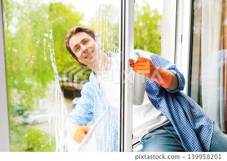 Smiling man doing spring cleaning and window washing at home. He wears orange gloves, using a spray bottle and squeegee for cleaning house glass. Smiling man doing spring cleaning and window washing at home. He wears orange gloves, using a spray bottle and squeegee for cleaning house glass. 139958201