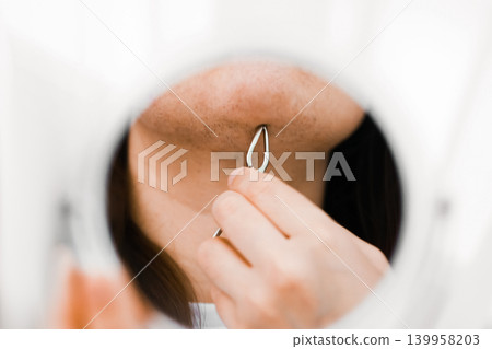 Reflection in a mirror of unwanted facial hair removal from the chin using a tool. Woman doing a home beauty routine for hair growth due to hormonal changes. Reflection in a mirror of unwanted facial hair removal from the chin using a tool. Woman doing a home beauty routine for hair growth due to hormonal changes. 139958203