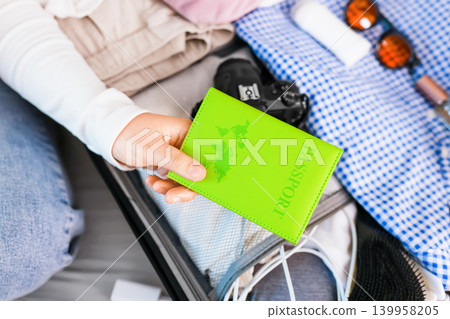 Hand holding a green passport over an open suitcase during travel preparation for a summer vacation. The luggage is packed with clothes and accessories. 139958205