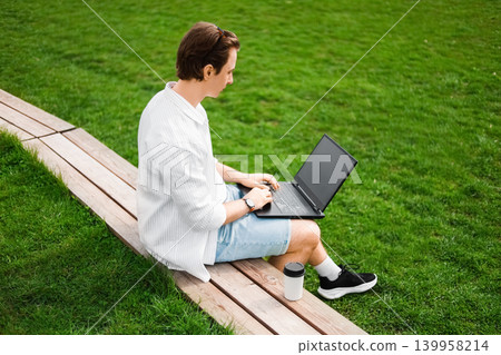 Freelancer doing remote work on a laptop outdoors. Man sitting on a wooden bench next to green grass, typing online with a coffee cup nearby. Freelancer doing remote work on a laptop outdoors. Man sitting on a wooden bench next to green grass, typing online with a coffee cup nearby. 139958214
