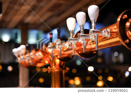 Close-up of polished copper beer taps with soft bokeh lights in a cozy bar setting. 139959081