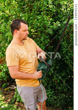 Person trimming garden hedge with electric hedge trimmer, backyard gardening maintenance 139959141