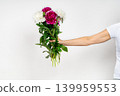 Close-up of a female hand holding and giving a beautiful bouquet of mixed white, pink, and burgundy peonies against a white background. 139959553