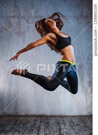 Young slim sports woman in black clothing jumping on gray wall background Young slim sports woman in black clothing jumping on gray wall background 139960109