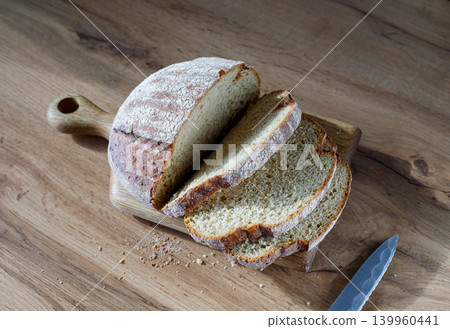 Sliced artisan wheat bread loaf with flour dusting on wooden cutting board, kitchen table meal preparation with sharp knife 139960441