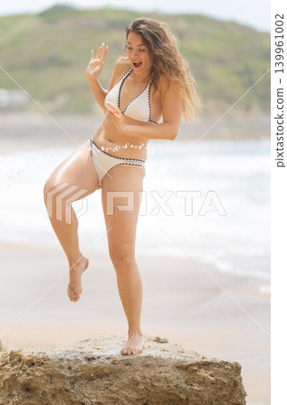 Woman has fun on a rock by the beach while splashing water with her foot during a sunny day at the shore Woman has fun on a rock by the beach while splashing water with her foot during a sunny day at the shore 139961002