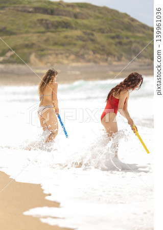 Two people play in the waves with colorful sticks on a sunny beach during the afternoon 139961036