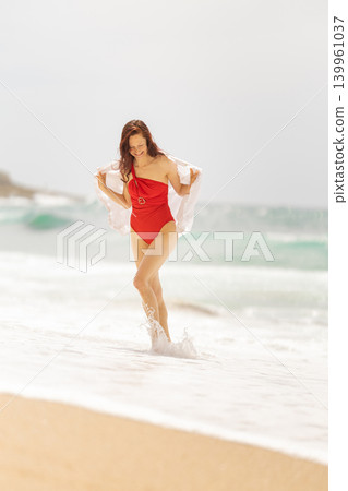 Woman walks along the beach in red swimsuit as waves crash on shore near calm sea during cloudy weather Woman walks along the beach in red swimsuit as waves crash on shore near calm sea during cloudy weather 139961037