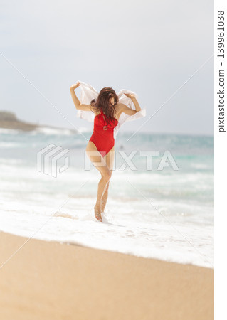 Woman walks on beach in red swimsuit holding white cloth by ocean shore during sunny day Woman walks on beach in red swimsuit holding white cloth by ocean shore during sunny day 139961038