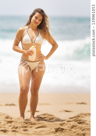 Woman stands on beach holding sunscreen bottle while ocean waves crash in the background during sunny day in summer Woman stands on beach holding sunscreen bottle while ocean waves crash in the background during sunny day in summer 139961053