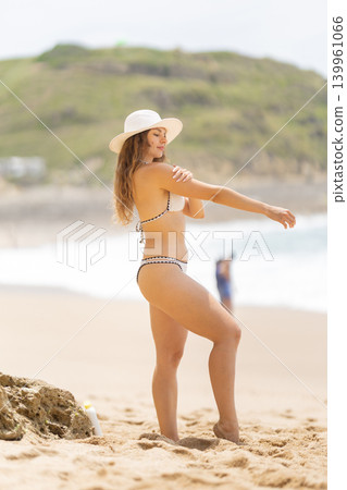 Woman in a bikini enjoys a sunny day at the beach while applying sunscreen near the ocean shore with other beachgoers around Woman in a bikini enjoys a sunny day at the beach while applying sunscreen near the ocean shore with other beachgoers around 139961066
