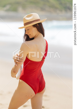 Woman in red swimsuit enjoys sunny beach day with waves in background and straw hat on head while standing on sandy shore 139961074