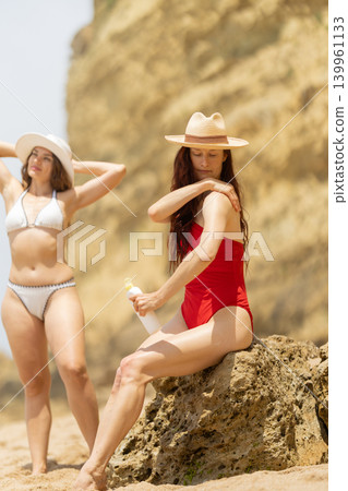 Women applying sunscreen at the beach during a sunny day with cliffs in the background and clear blue sky Women applying sunscreen at the beach during a sunny day with cliffs in the background and clear blue sky 139961133