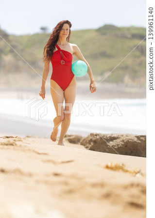 Woman walks along the beach carrying a blue ball on a sunny day near the shoreline with green hills in the background 139961140