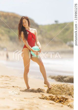 Woman stands on sandy beach holding a blue ball with waves in background during sunny day near rocky shore 139961276