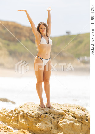 Female model in white bikini poses on a rock at the beach, arms raised, with ocean waves and green hills in the background under a bright sky Female model in white bikini poses on a rock at the beach, arms raised, with ocean waves and green hills in the background under a bright sky 139961292