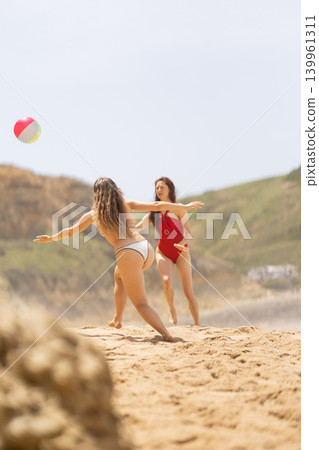 Two women playing with a beach ball on sandy shore, one in red swimsuit and the other in white bikini, with green hills and blue sky in the background Two women playing with a beach ball on sandy shore, one in red swimsuit and the other in white bikini, with green hills and blue sky in the background 139961311