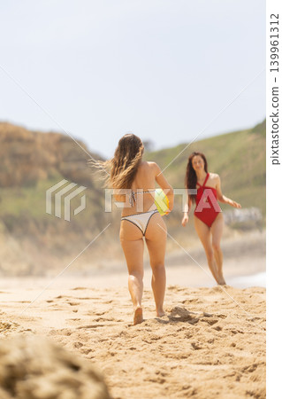 Two women enjoying a sunny day at the beach, one in a black and white bikini carrying a yellow ball, the other in a red swimsuit standing on the sandy shore Two women enjoying a sunny day at the beach, one in a black and white bikini carrying a yellow ball, the other in a red swimsuit standing on the sandy shore 139961312