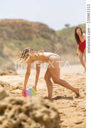 Young woman in a bikini playing with a colorful beach ball on sandy shore, with a rocky landscape and another person in a red swimsuit in the background Young woman in a bikini playing with a colorful beach ball on sandy shore, with a rocky landscape and another person in a red swimsuit in the background 139961313