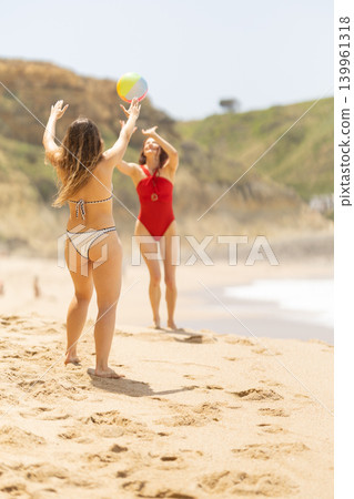 Two women playing with a beach ball on sandy shore, one in a red swimsuit and the other in a bikini, with ocean waves and green hills in the background 139961318
