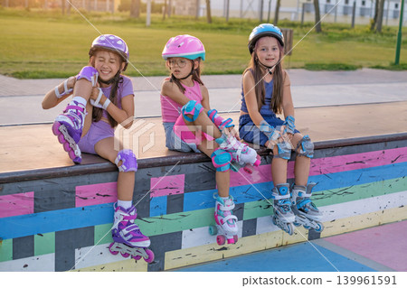 Three cheerful tired girlfriends are sitting on a ramp in a skatepark in sports protective gear and roller skates Three cheerful tired girlfriends are sitting on a ramp in a skatepark in sports protective gear and roller skates 139961591