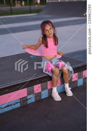 A girl is sitting on the ramp of a skate park holding a pink skateboard in her hands. Playing sports in the summer until the evening A girl is sitting on the ramp of a skate park holding a pink skateboard in her hands. Playing sports in the summer until the evening 139961593
