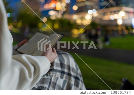 A person opening a book outdoors at night, with the beautiful city lights in bokeh. A person opening a book outdoors at night, with the beautiful city lights in bokeh. 139961724