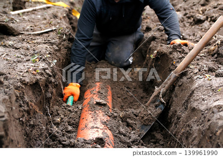 Worker laying an orange PVC drainage pipe in a trench. Process of installing an underground sewer or water system in the ground with a shovel and manual labor Worker laying an orange PVC drainage pipe in a trench. Process of installing an underground sewer or water system in the ground with a shovel and manual labor 139961990