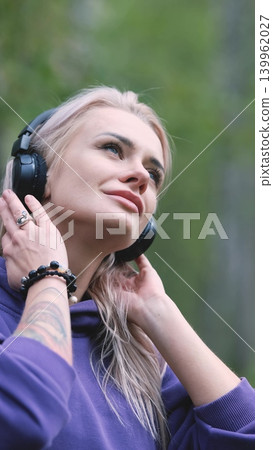 Blonde in purple sweatshirt listening to music on headphones while breathing fresh air in forest Blonde in purple sweatshirt listening to music on headphones while breathing fresh air in forest 139962027