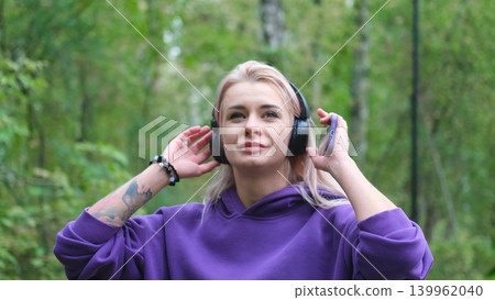 Blonde in purple sweatshirt listening to music on headphones while breathing fresh air in forest Blonde in purple sweatshirt listening to music on headphones while breathing fresh air in forest 139962040