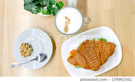 Flat lay top view of a savory croissant sandwich served with a hot coffee latte, a small shortbread cookie, and a green potted plant on a bright wooden cafe table Flat lay top view of a savory croissant sandwich served with a hot coffee latte, a small shortbread cookie, and a green potted plant on a bright wooden cafe table 139962562